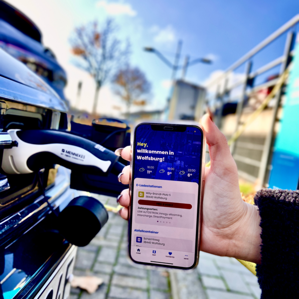 Hand hält Handy mit WolfsburgApp in der Hand. Im Hintergrund ein ladendes Auto.