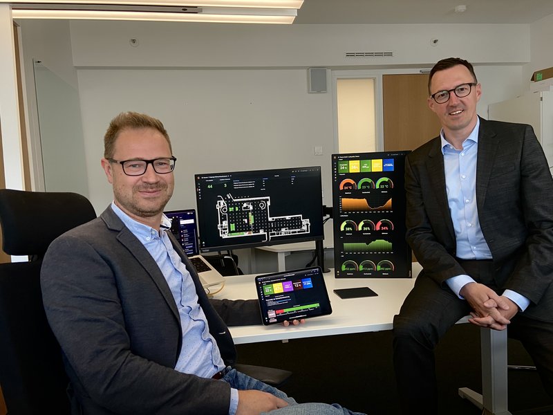 Anatoli Seliwanow, Leiter Betrieb bei der Wobcom GmbH (rechts) und Dalibor Dreznjak, Leiter Unternehmensentwicklung und Kommunikation, Stadtwerke Wolfsburg AG, sind stolz auf die Plattform-Lösung der Stadtwerke Wolfsburg. Im Hintergrund sind verschiedene Dashboard-Ansichten zu sehen, wie Außen- und Innenluft, Parken in der Tiefgarage. 