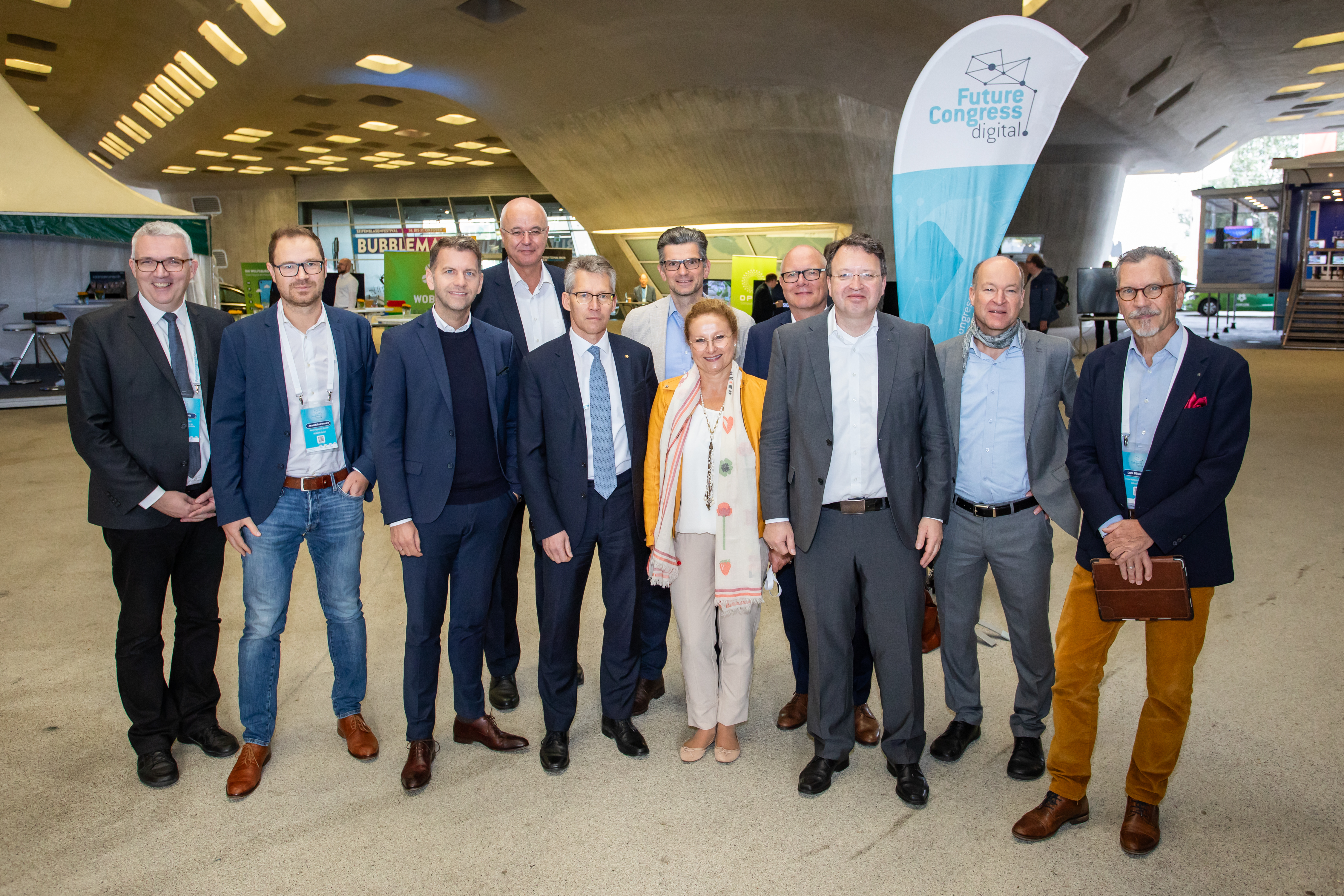 Die Teilenehmenden des Future-Congress.digital stehen nebeneinander aufgereit unter dem phaeno Wolfsburg und lächeln in die Kamera. Im Hintergrund steht eine Beachflag mit dem Logo des Kongresses.