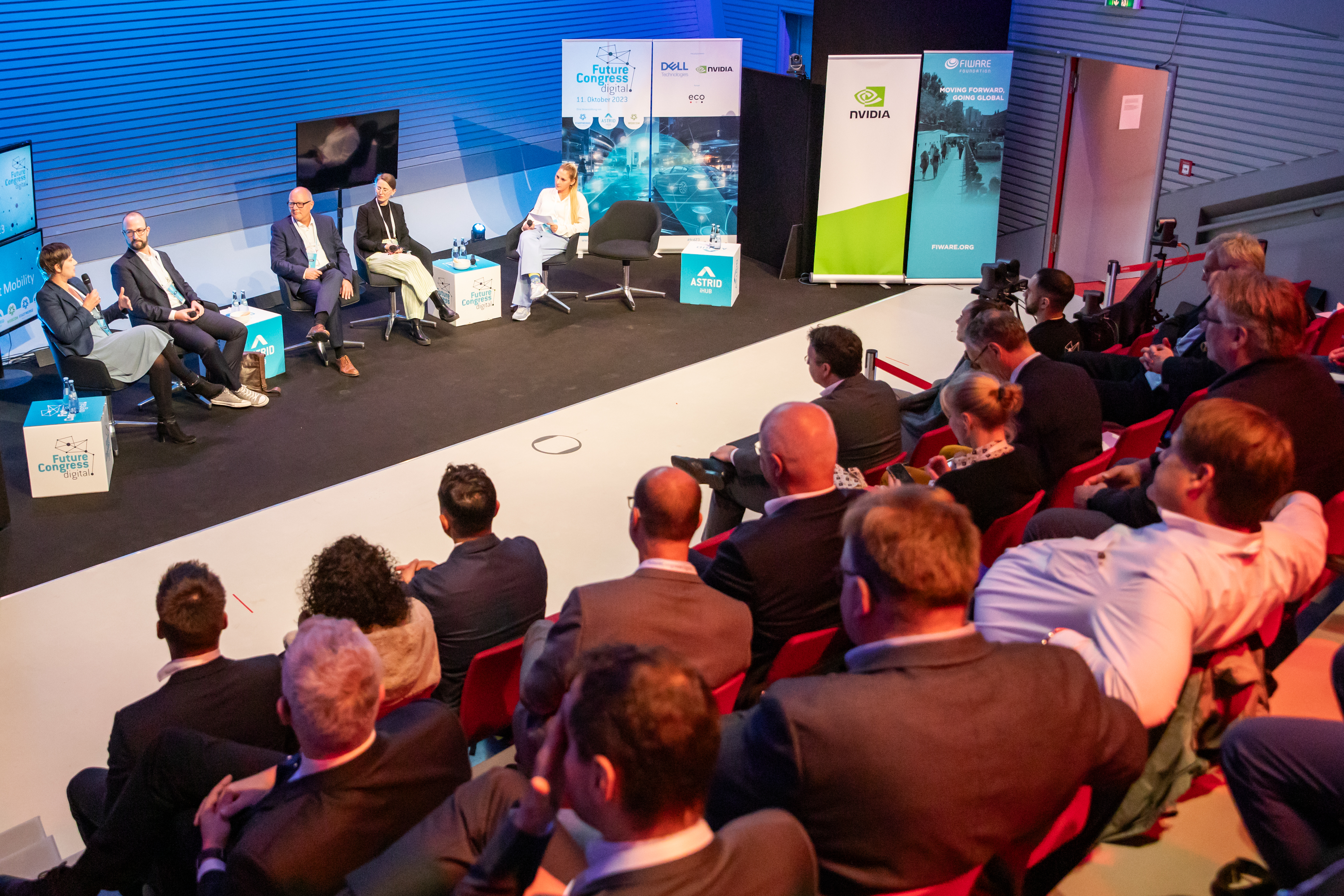 Menschen auf der Tribüne hören und schauen der Paneldiskussion unten auf der Bühne zu.