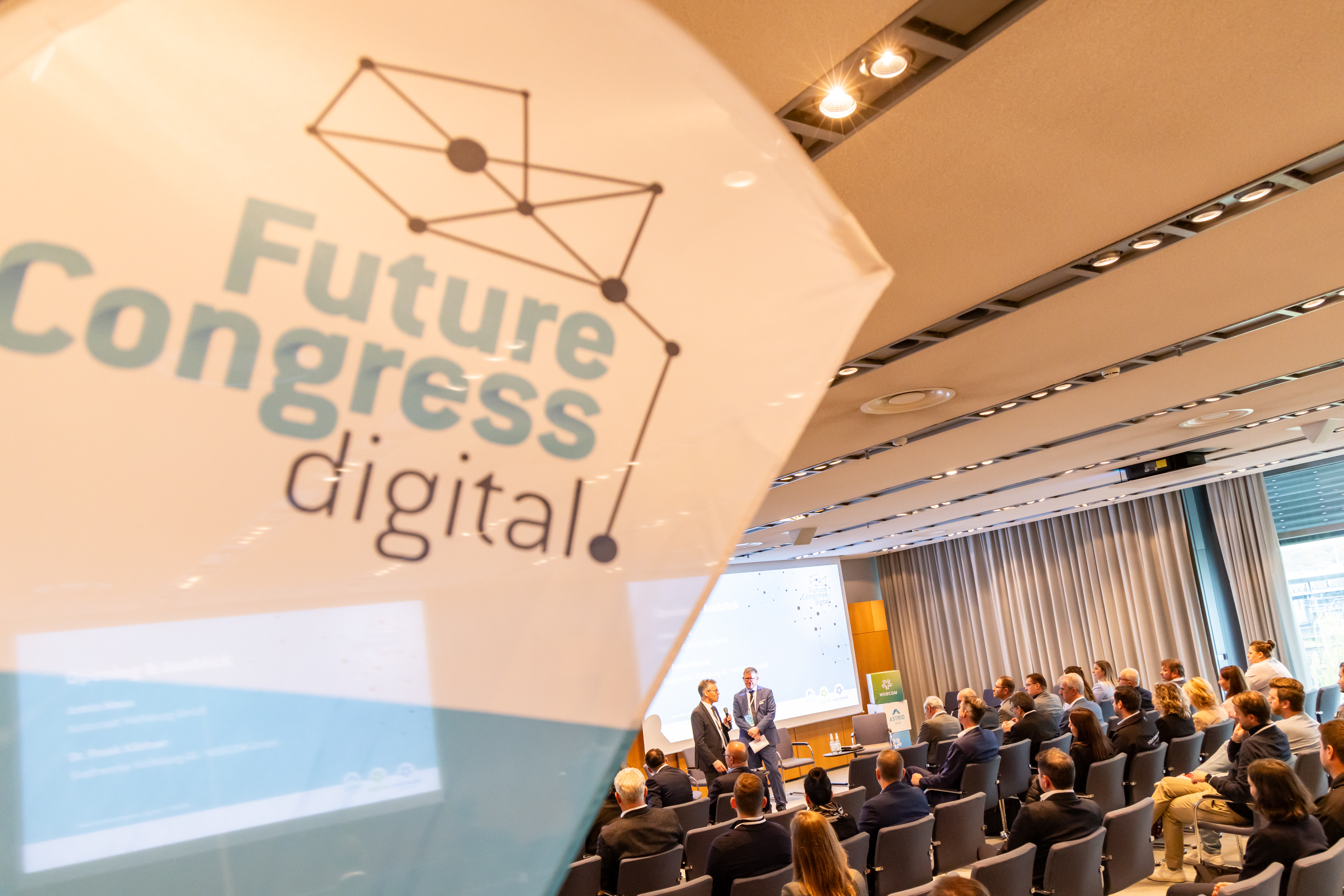 Das FutureCongress.digital Logo im Vordergrund. Im Hintergrund sieht man das Plenum und Dr. Frank Kästner zusammen mit Armin Maus bei der Begrüßung