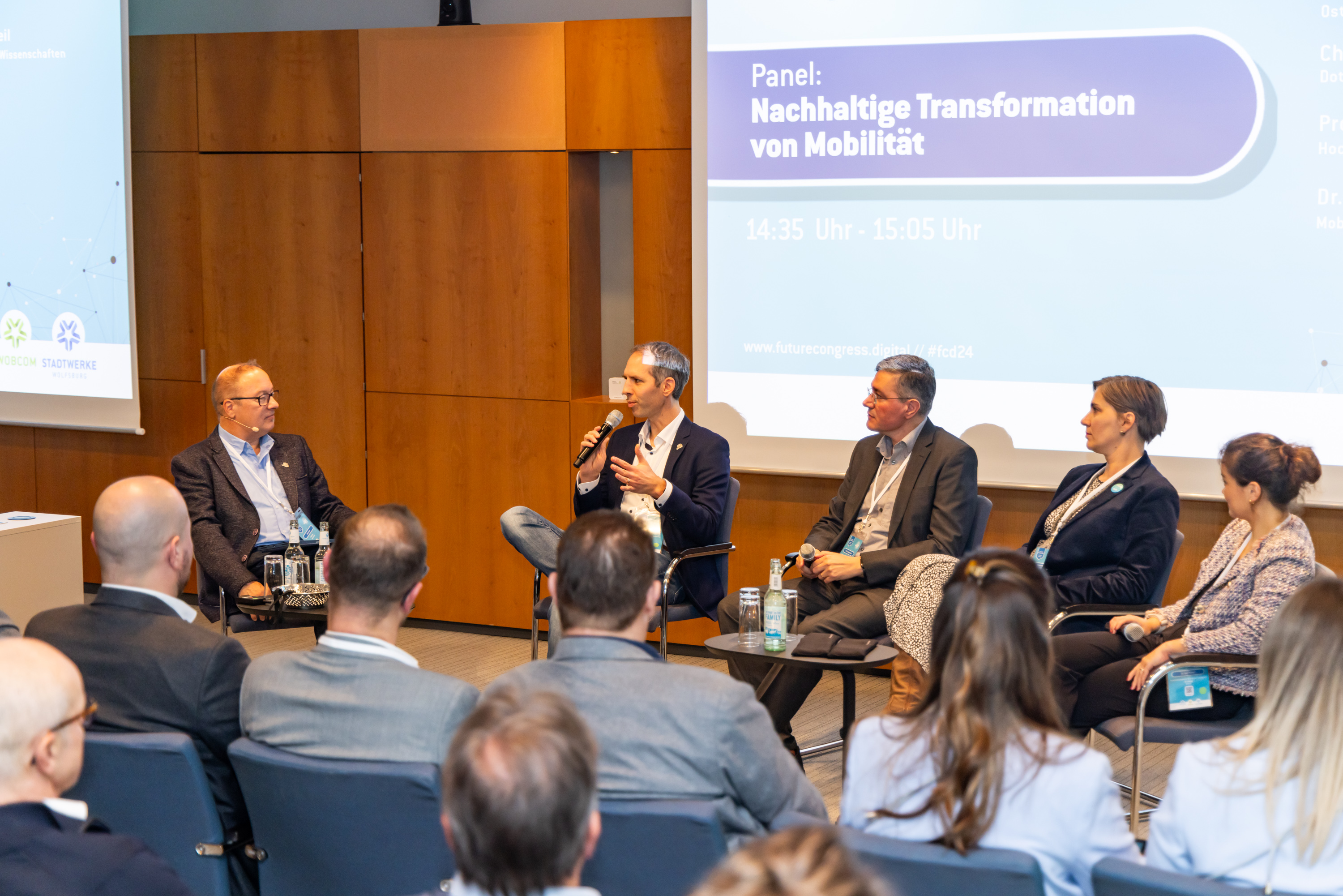 Drei Männer und zwei Frauen sitzen im Halbkreis vor der Leinwand und diskutieren über das Thema: Nachhaltige Transformation von Mobilität