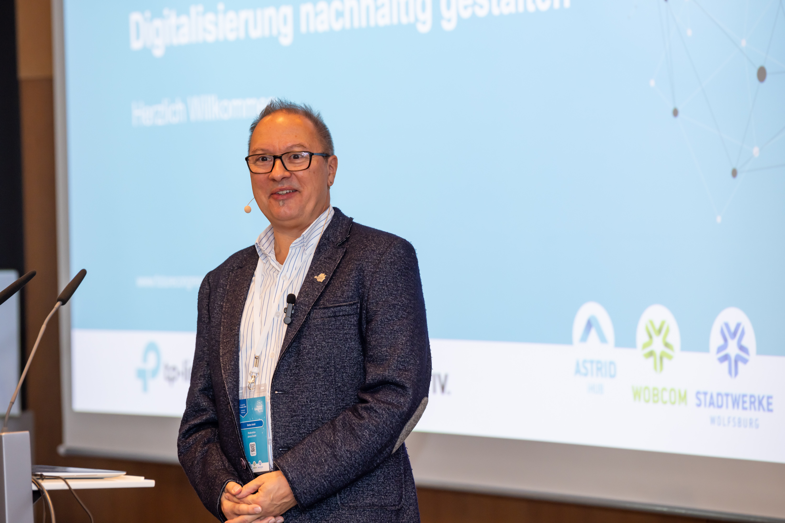 Der Moderator Detlev Artelt steht vorne am Rednerpult vor der Leinwand und eröffnet den Kongress